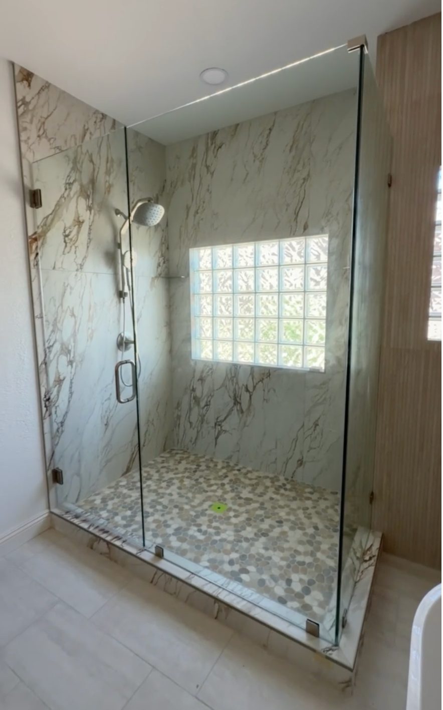 A modern walk-in shower enclosure with a frameless glass door, featuring a marble-look tiled wall and floor, a glass block window, and a chrome handheld shower fixture.