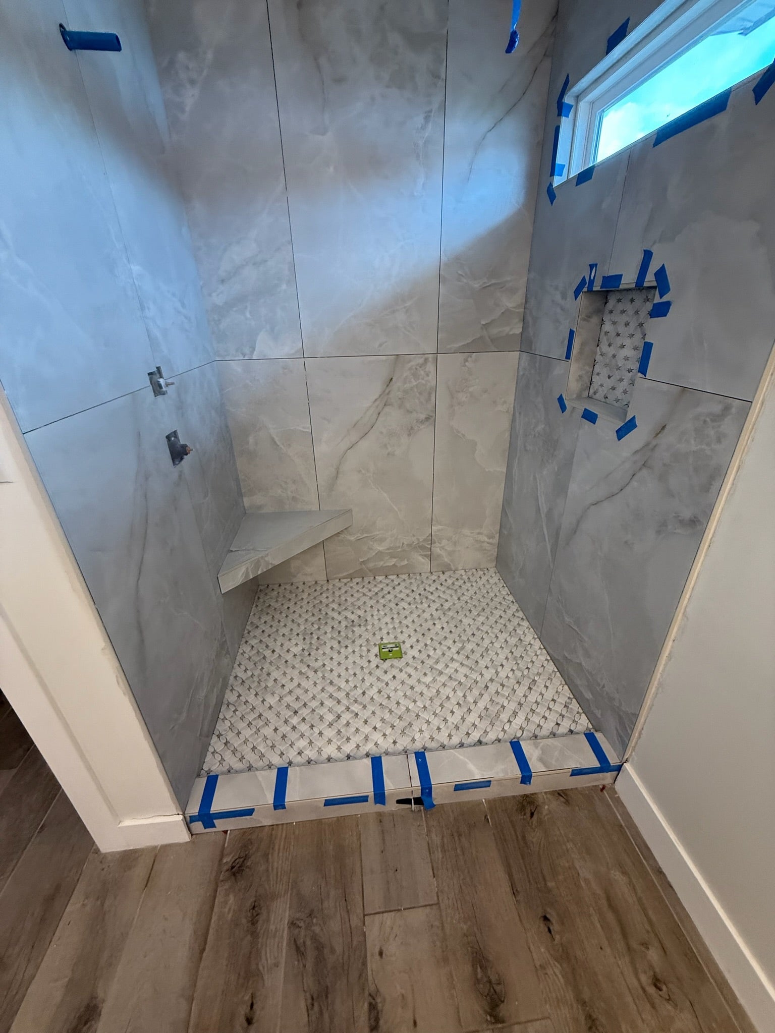 A walk-in shower stall with large light gray marble-look tiles on the walls, a white mosaic tile floor, a built-in bench, a small niche outlined with blue painter's tape, and wood-look flooring around the shower base.