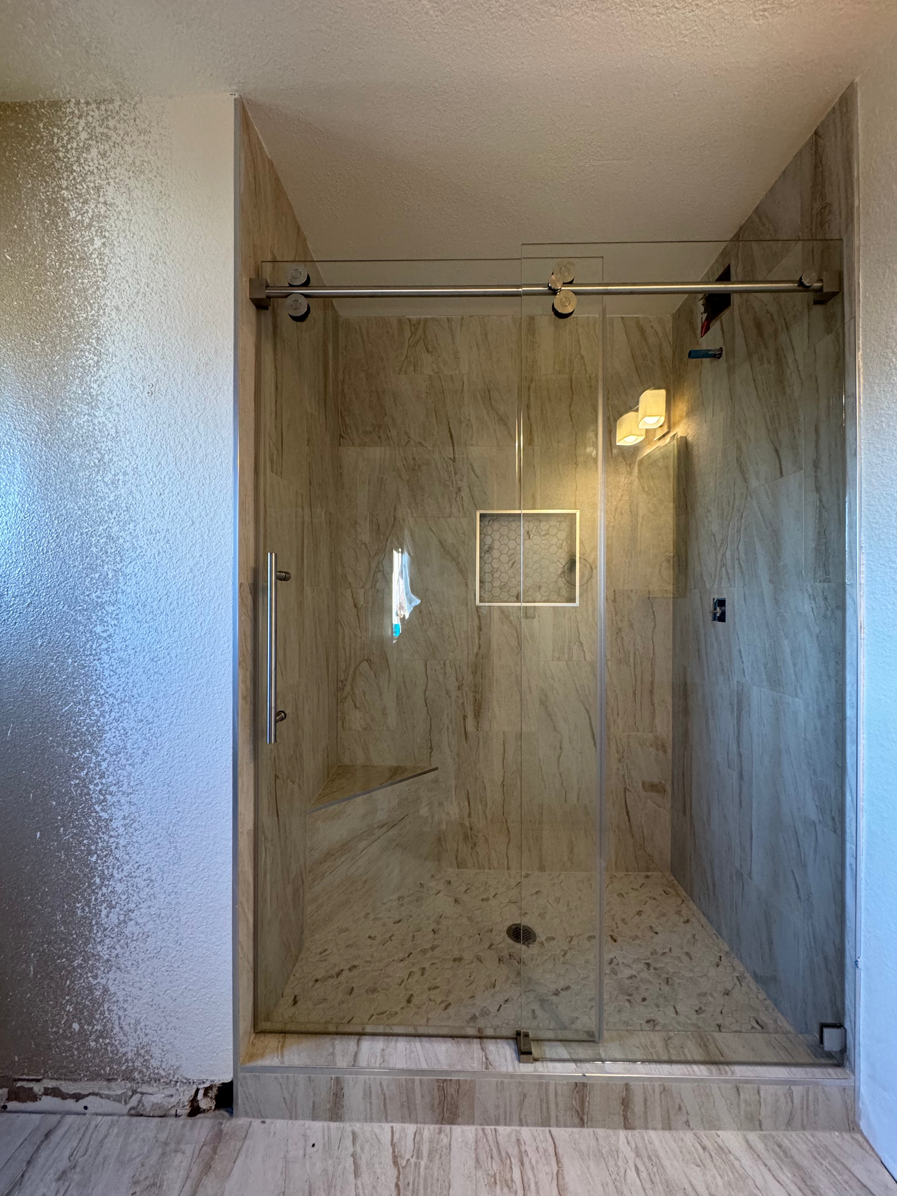 A modern walk-in shower enclosure with a clear glass door and a sliding barn-style door, set in a bathroom with beige marble-look tile walls and floor.