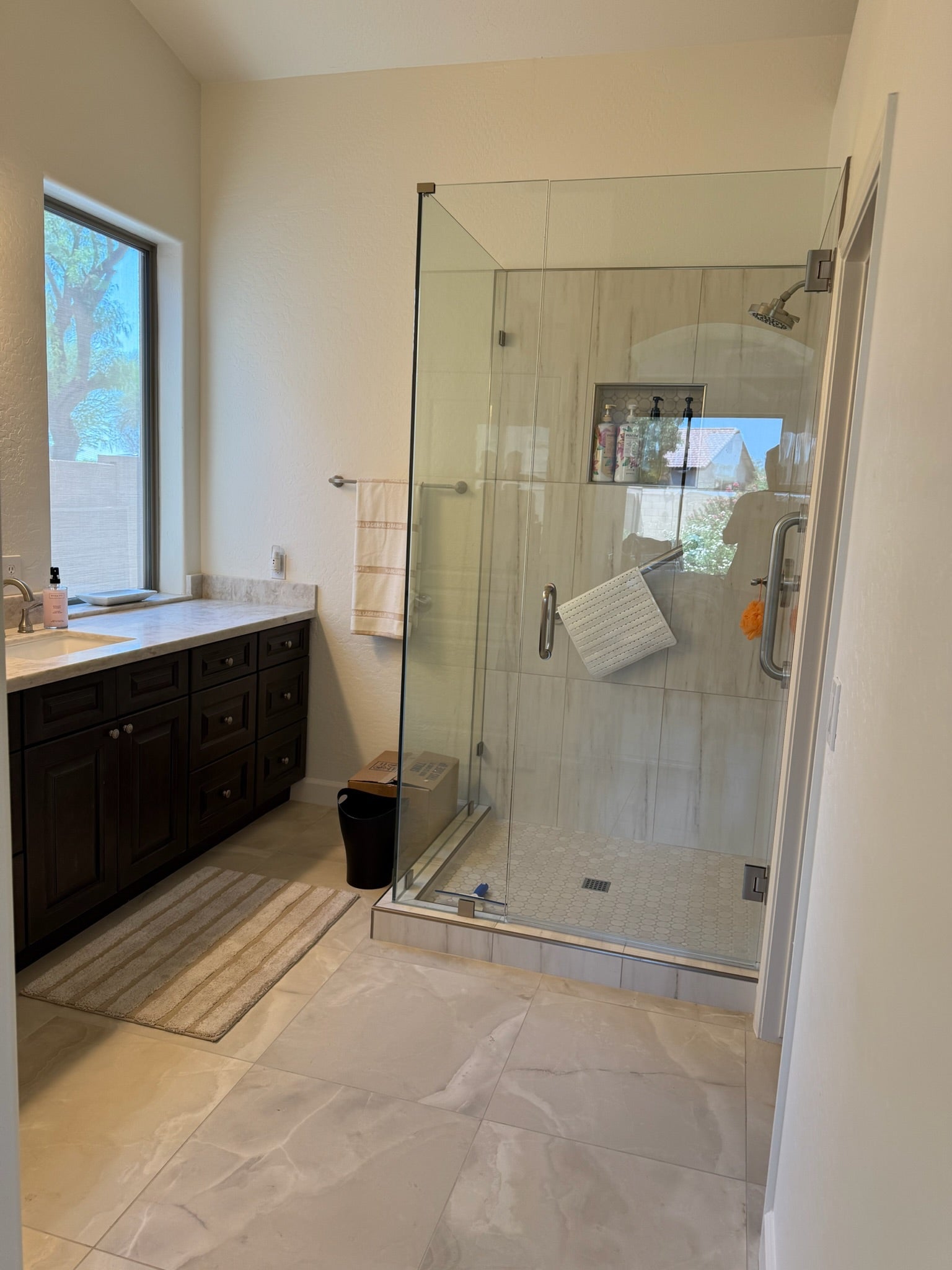 A modern bathroom featuring a walk-in glass shower enclosure with a tiled shower floor, a dark wood vanity with a white countertop, and light beige stone tile flooring.